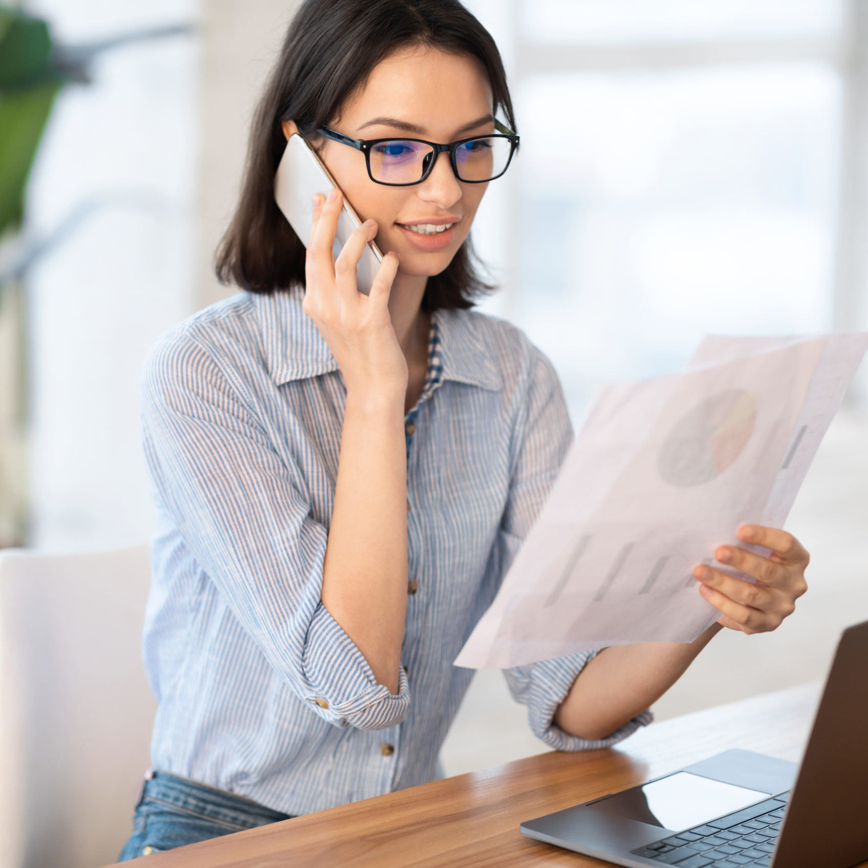 Corporate Communication. Young business woman in glasses making phone call, holding papers with graphs, copyspace